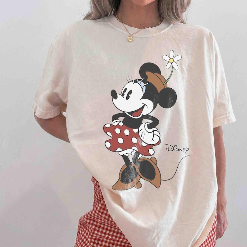 May include: Cream-colored t-shirt featuring a Minnie Mouse graphic. Minnie is depicted in a red polka-dot dress, brown shoes, and a hat with a flower. The Disney logo is printed on the shirt.