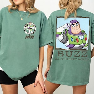 Puede incluir: Una camiseta verde salvia con un gr&aacute;fico de Buzz Lightyear en la espalda y un gr&aacute;fico m&aacute;s peque&ntilde;o de Buzz Lightyear en la parte delantera. El gr&aacute;fico frontal est&aacute; dentro de un c&iacute;rculo a cuadros con el nombre "ANDY". El gr&aacute;fico trasero dice "BUZZ WALT DISNEY WORLD".
