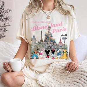 May include: Cream-colored t-shirt featuring a graphic of Disneyland Paris with Mickey Mouse, Minnie Mouse, Donald Duck, and other Disney characters. The text "Paris, France Est 1992 Disneyland Paris" is printed in pink.