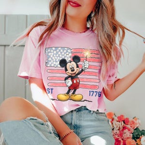 May include: Pink t-shirt featuring Mickey Mouse holding a sparkler in front of an American flag design. The shirt has the text "1776" and "ST" printed on it. The shirt is a light pink color.