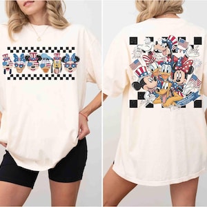 May include: Cream-colored t-shirt featuring a patriotic design with cartoon characters. The front displays a row of character faces above a black and white checkered pattern. The back showcases a group of characters with American flags and fireworks.
