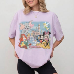 May include: A light purple t-shirt featuring a Disneyland graphic. The graphic includes various Disney characters such as Dumbo, Ariel, Cinderella, Mickey and Minnie Mouse, and Tinkerbell, with the Disneyland castle in the background.