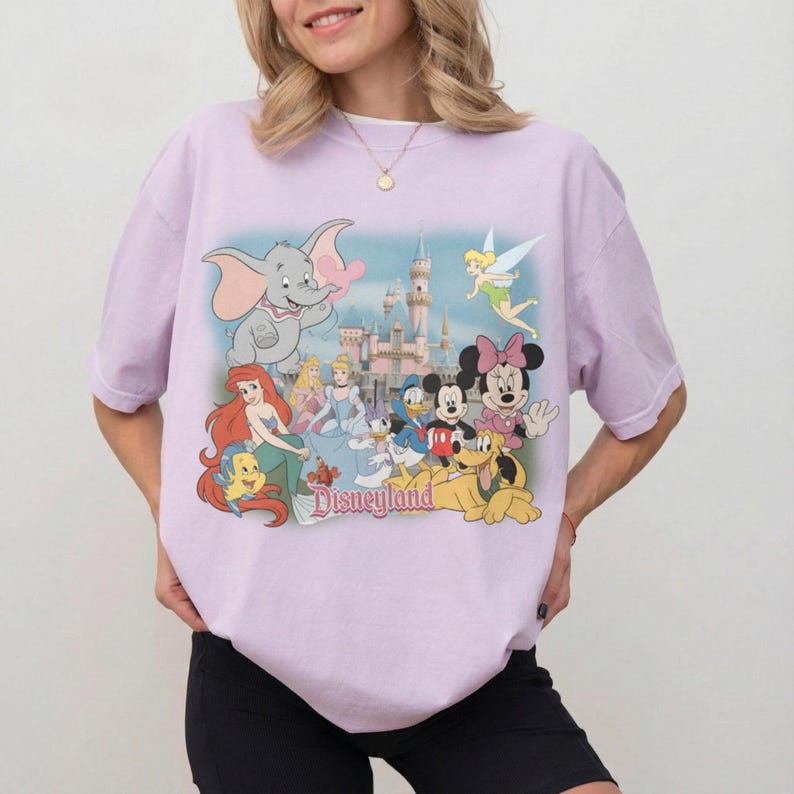 May include: A light purple t-shirt featuring a Disneyland graphic. The graphic includes various Disney characters such as Dumbo, Ariel, Cinderella, Mickey and Minnie Mouse, and Tinkerbell, with the Disneyland castle in the background.