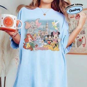 May include: Light blue t-shirt featuring a Disneyland graphic with various cartoon characters, including Dumbo, Mickey and Minnie Mouse, and Ariel. The shirt has a relaxed fit and is made of a soft material. A white mug with a sun design is held in the hand.