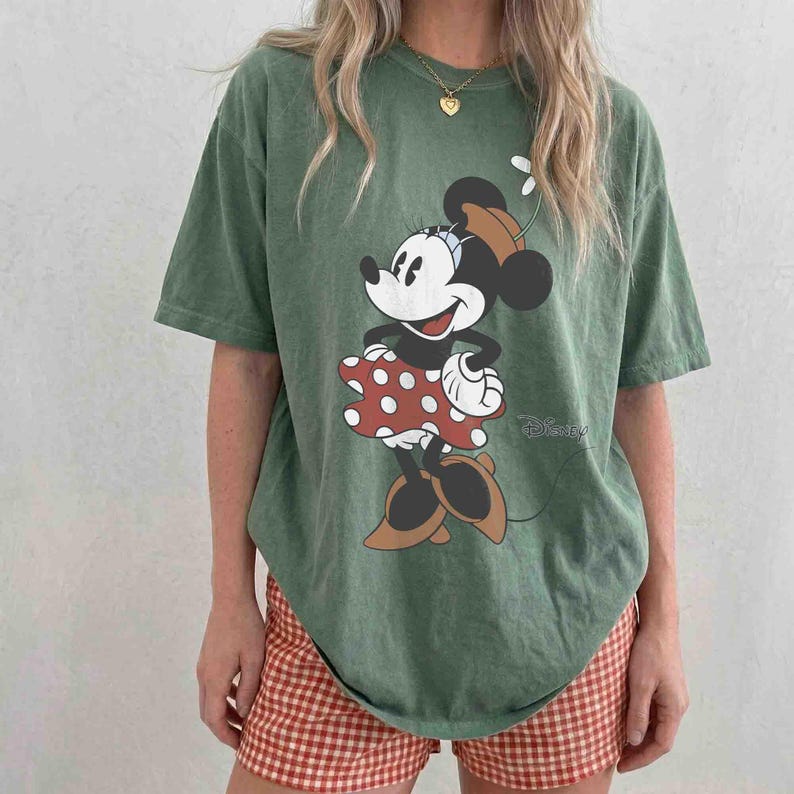 May include: A green t-shirt featuring a Minnie Mouse graphic. The cartoon character is wearing a red polka dot dress and brown shoes. The shirt has a relaxed fit and is paired with red and white checkered shorts.