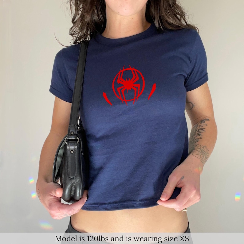 Puede incluir: Camiseta azul marino con un gr&aacute;fico de ara&ntilde;a roja en la parte delantera. La camiseta de manga corta es de estilo corto. La modelo lleva la talla XS.