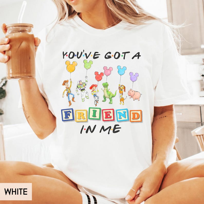 May include: A white t-shirt featuring a graphic of Toy Story characters holding Mickey Mouse-shaped balloons. The text reads "YOU'VE GOT A FRIEND IN ME" with the word "FRIEND" displayed on colorful alphabet blocks. The design includes Woody, Buzz Lightyear, Jessie, Rex, Slinky Dog, and Hamm.
