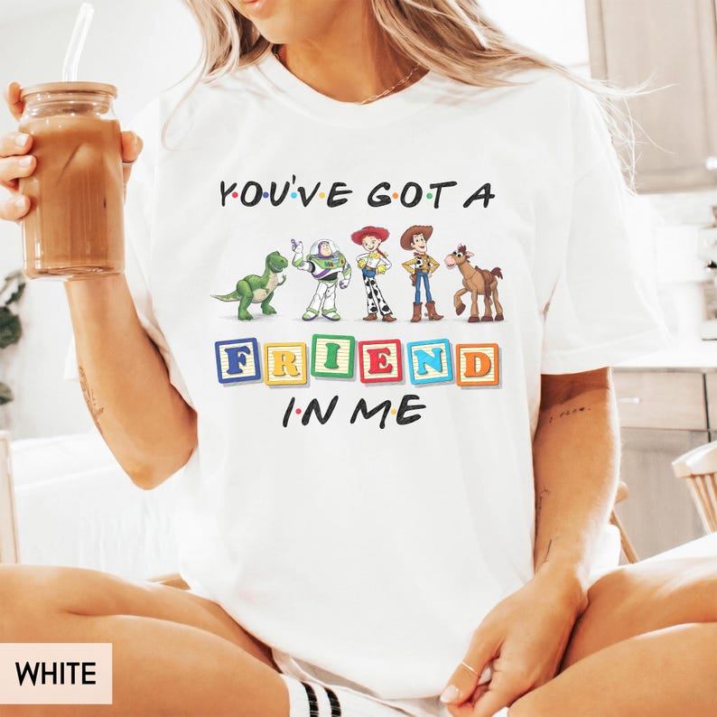 May include: A white short-sleeve t-shirt featuring a graphic print of Toy Story characters including Rex, Buzz Lightyear, Jessie, Woody, and Bullseye. The text reads You've Got A Friend In Me with the word Friend spelled out in colorful alphabet blocks.