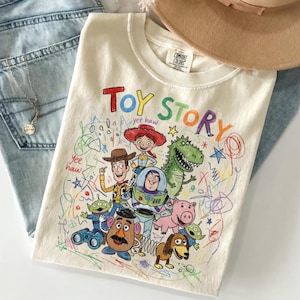 May include: Cream-colored t-shirt featuring a colorful "Toy Story" graphic with Woody, Buzz Lightyear, and other characters. The design includes the text "Toy Story" in a playful font, with the words "yee haw" written in the design.