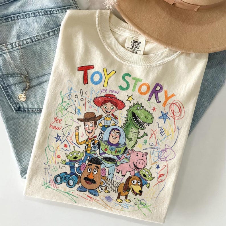 May include: Cream-colored t-shirt featuring a colorful "Toy Story" graphic with Woody, Buzz Lightyear, and other characters. The design includes the text "Toy Story" in a playful font, with the words "yee haw" written in the design.