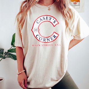May include: Cream-colored t-shirt featuring the text "Casey's Corner" in a red and blue design. The shirt also includes the text "Main Street, USA" in red. The design is centered on the front of the shirt.