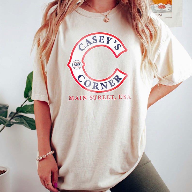 May include: Cream-colored t-shirt featuring the text "Casey's Corner" in a red and blue design. The shirt also includes the text "Main Street, USA" in red. The design is centered on the front of the shirt.