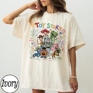 May include: Ivory-colored t-shirt featuring a colorful graphic of Toy Story characters. The design includes Woody, Buzz Lightyear, and other characters from the animated film, with the words "Toy Story" in a playful font. The shirt has a relaxed fit.