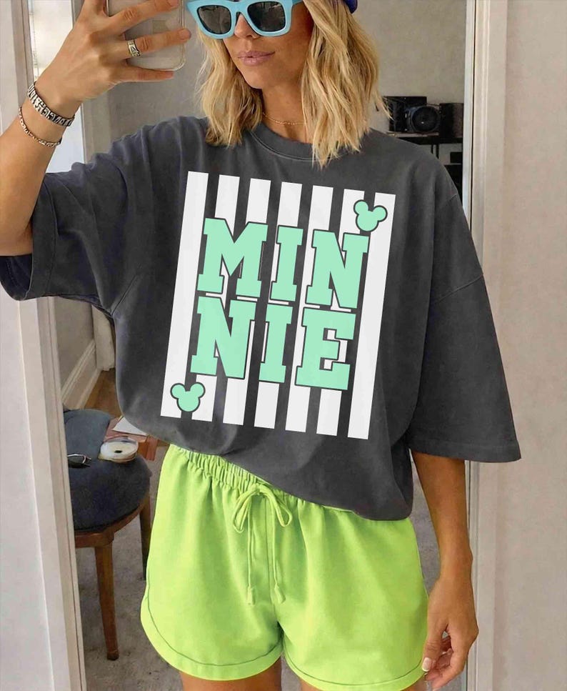 May include: Dark gray t-shirt with the word "MINNIE" in mint green letters, framed by white stripes. The shirt is paired with neon green shorts. The outfit is casual and comfortable.