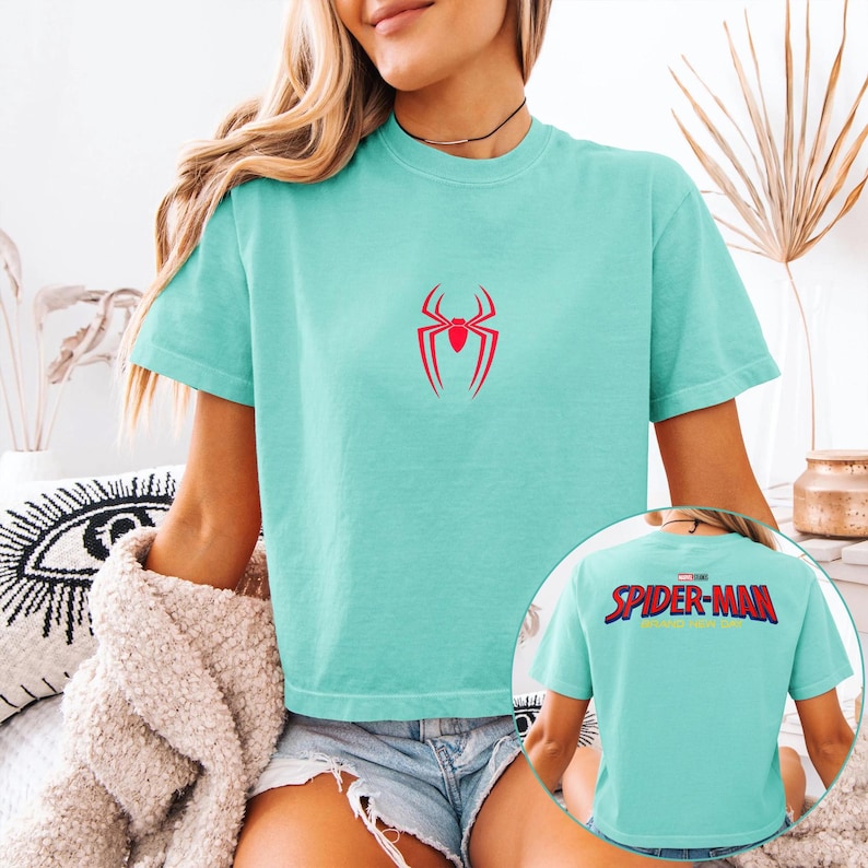 May include: A light teal t-shirt featuring a red spider emblem on the front. The back of the shirt displays the text "SPIDER-MAN" in a colorful, block letter design. The shirt is short-sleeved and has a relaxed fit.