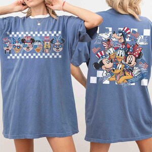 May include: Blue t-shirt featuring a graphic of Disney characters in patriotic attire, including Mickey Mouse, Minnie Mouse, and Donald Duck. The front design is above a white and black checkered pattern. The back of the shirt also features a similar design.