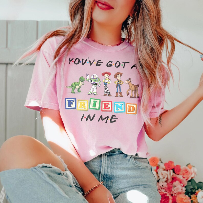 May include: A light pink short-sleeve t-shirt featuring a graphic of Toy Story characters including Rex, Buzz Lightyear, Jessie, Woody, and Bullseye. The text reads You've Got A Friend In Me with the word Friend spelled out in colorful toy blocks.