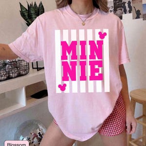 May include: A light pink t-shirt with the word "MINNIE" in bright pink block letters, set against a white striped background. The shirt features two small pink Mickey Mouse silhouettes. The shirt is part of a casual wear collection.