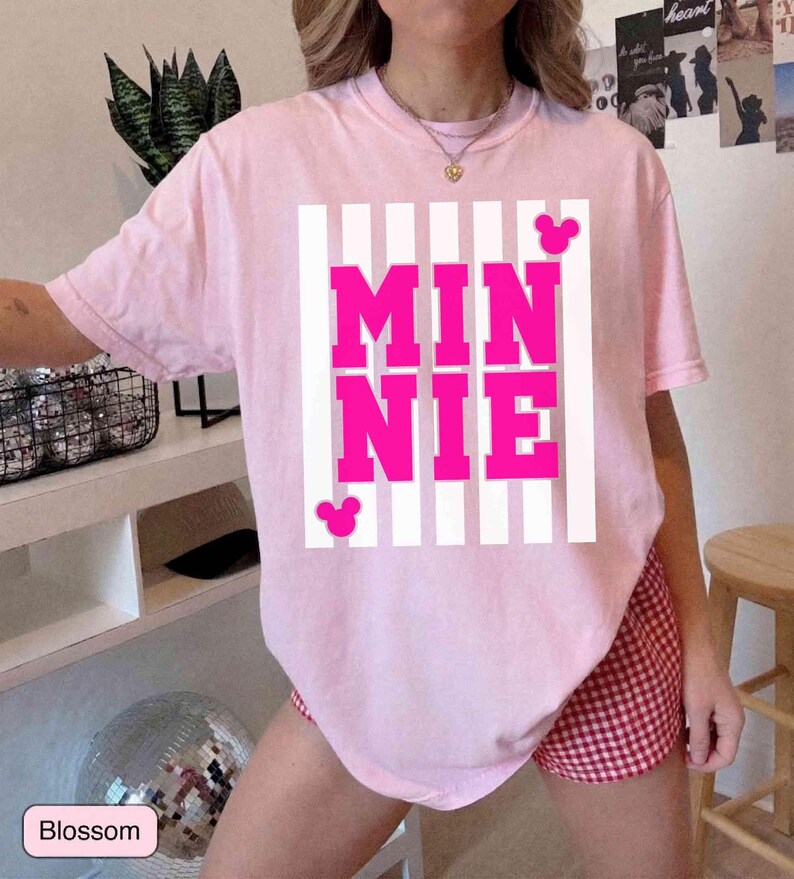 May include: A light pink t-shirt with the word "MINNIE" in bright pink block letters, set against a white striped background. The shirt features two small pink Mickey Mouse silhouettes. The shirt is part of a casual wear collection.