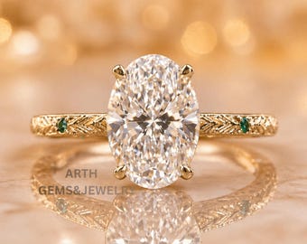 Anillo de compromiso ovalado con diamante creado en laboratorio, detalles de esmeralda en oro amarillo y banda grabada.