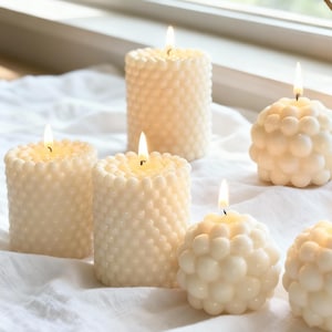 May include: Several ivory-colored candles with lit flames are arranged on a white surface. The candles have a textured, bubble-like design. Some are cylindrical, while others are spherical, creating a warm and inviting ambiance.