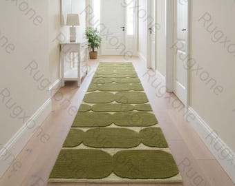Puede incluir: Una alfombra larga de color verde oliva con un patrón repetitivo de formas redondeadas, colocada sobre un suelo de madera clara. La alfombra está en un pasillo con paredes y puertas blancas.