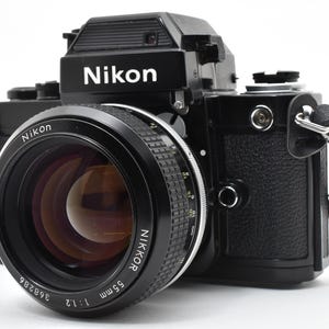 May include: A black Nikon SLR film camera featuring a Nikkor 55mm 1:1.2 lens. The camera body has a textured grip, a pentaprism housing with the Nikon logo, and a black fabric neck strap attached to the side lugs. The lens barrel displays white text.