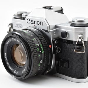 May include: A silver and black Canon AE-1 35mm film camera featuring a Canon Lens FD 50mm 1:1.8 lens. The camera body has a textured black grip, a silver top plate with the Canon logo, and a lens barrel with green and white markings.
