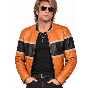 Puede incluir: Una chaqueta de cuero color canela con una franja horizontal negra en el pecho y los brazos, bordeada por finas líneas beige. La chaqueta tiene cuello redondo, cierre de cremallera frontal y mangas largas con puños de cremallera sobre camiseta.