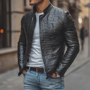 May include: A black leather jacket with a crocodile skin effect. The jacket features a stand-up collar and a zip closure. The person is wearing a white t-shirt and blue jeans. A brown belt and a watch are also visible.