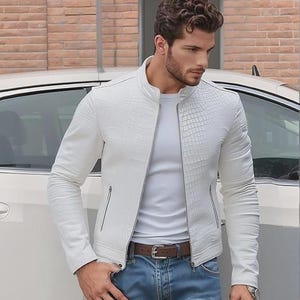 Peut inclure: Veste blanche en similicuir avec un motif texturé en forme de crocodile sur les épaules et le haut des bras. La veste a une fermeture éclair, deux poches latérales et un col montant. Le modèle porte un t-shirt blanc, un jean bleu et une ceinture marron.