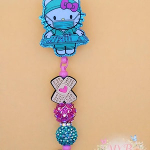May include: A badge reel featuring a cartoon character in medical attire. The design includes a Hello Kitty character wearing a surgical cap and mask, a bandage, and two decorative beaded spheres. The reel is attached to a clear plastic clip.