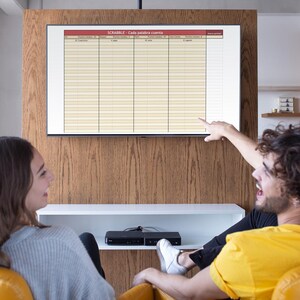 May include: A television screen displays a Scrabble game sheet with columns for words and scores. Two people are seated in front of the screen, one pointing at the game. The room has a wooden accent wall and a white shelf.