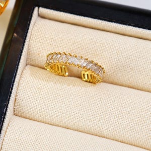 May include: A gold-toned open ring featuring a row of clear, rectangular baguette-cut stones set along the top band. The metal band has a textured, ribbed design on the sides and an open back, allowing for adjustable sizing on the finger.