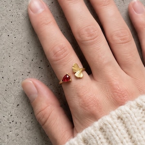 Peut inclure: Une bague ouverte en or ornée d'une petite pierre rouge en forme de goutte d'eau à une extrémité et d'un motif de feuille de ginkgo en éventail texturé à l'autre. La bague est portée au doigt, mettant en valeur la pierre rouge et la feuille.