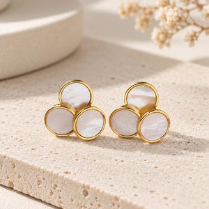 May include: A pair of gold-toned stud earrings, each featuring three circular white mother-of-pearl inlays arranged in a clover-like cluster. The metal settings are polished gold, framing the iridescent, flat, round shell discs securely.