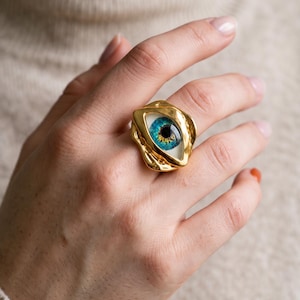 May include: A gold-toned ring featuring an eye motif with a realistic blue iris and black pupil set within an irregular, organic-shaped metal frame. The ring has a polished, reflective surface and a smooth, rounded band design.
