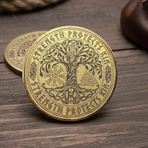 Può includere: Due monete color oro con disegni intricati e l'iscrizione "STRENGTH PROTECTS KIN" sul bordo. Le monete presentano un simbolo dell'albero della vita e altre immagini di ispirazione norrena. Lo sfondo include corda e pelle.
