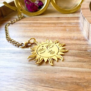 May include: A gold-colored tea infuser with a sun-shaped charm. The charm has a face with closed eyes and star details. The infuser is attached to a chain and is resting on a wooden tray. The infuser is filled with dried rose buds.
