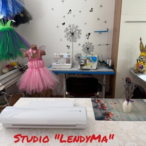 Op de afbeelding: Een naaiatelier met een roze tutu-jurk op een mannequin. De studio beschikt over naaimachines, een witte tafel met een snijmachine en een muur versierd met paardenbloem- en vlinderstickers. De tekst "STUDIO "LENDYMA"" is zichtbaar.