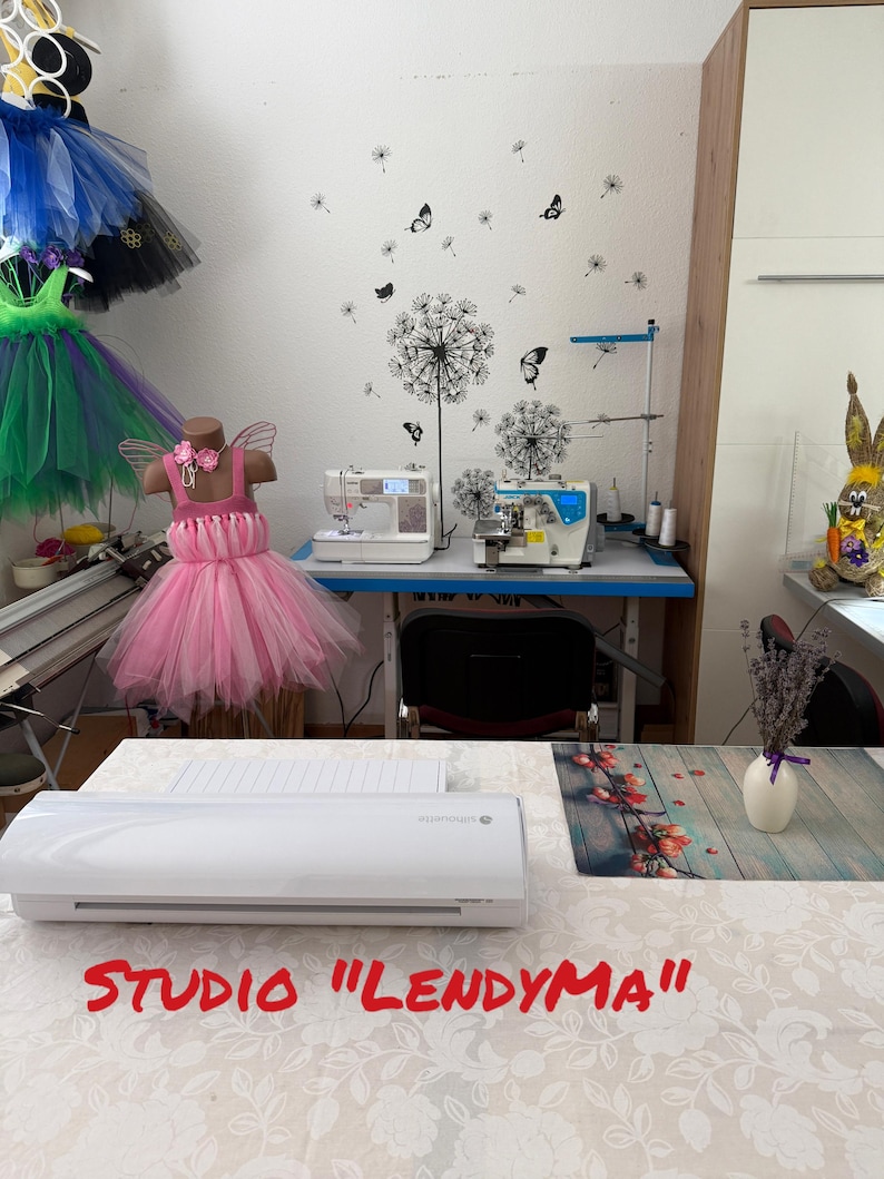 Op de afbeelding: Een naaiatelier met een roze tutu-jurk op een mannequin. De studio beschikt over naaimachines, een witte tafel met een snijmachine en een muur versierd met paardenbloem- en vlinderstickers. De tekst "STUDIO "LENDYMA"" is zichtbaar.