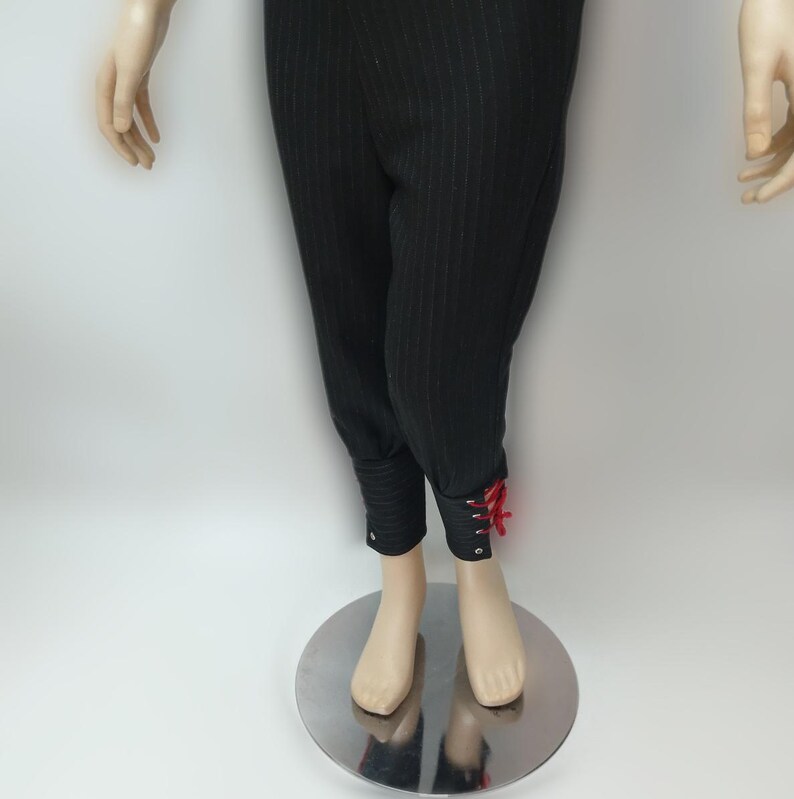 Op de afbeelding: Zwarte broek met krijtstreep en rode veterdetails bij de enkels. De broek heeft een getailleerde, cropped stijl en wordt op een mannequin getoond. De stof lijkt een gebreid of geweven materiaal te zijn.