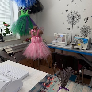 May include: A pink tutu dress with a bodice and tulle skirt is displayed on a mannequin. Other colorful tutus hang in the background. Sewing machines and a work table are also visible, suggesting a handmade clothing business.