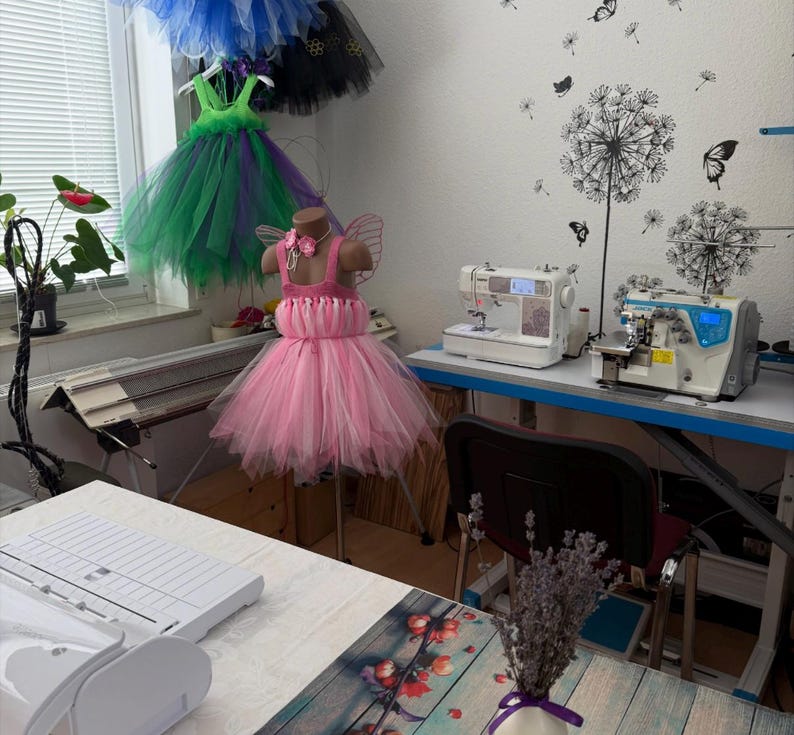 May include: A pink tutu dress with a bodice and tulle skirt is displayed on a mannequin. Other colorful tutus hang in the background. Sewing machines and a work table are also visible, suggesting a handmade clothing business.