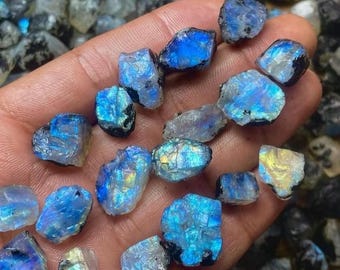 May include: A collection of raw, uncut moonstone gemstones. The stones display a range of colors, including blues, whites, and hints of yellow. The gemstones are held in a hand, showcasing their natural, irregular shapes and textures. The background is filled with more moonstones.