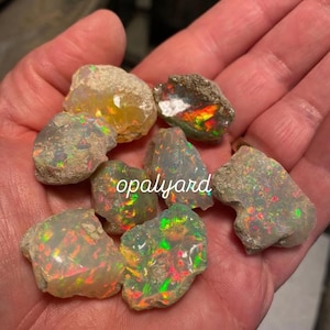 Puede incluir: Una colección de piedras de ópalo en bruto, que muestran una variedad de colores, incluyendo verde, rojo y amarillo. Los ópalos tienen un aspecto natural, sin pulir, con texturas y patrones visibles. La palabra "opalyard" está escrita en las piedras.