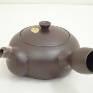 May include: A brown ceramic teapot with a rounded body, a lid with a small knob, and a side handle. The spout is angled and the handle is a large, open cylinder. A small, round sticker is on the lid.
