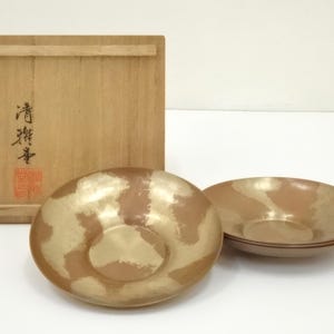 Op de afbeelding: Twee bronskleurige kommen en een houten kist. De kommen hebben een cirkelvorm met een verhoogd midden en een metallic goud- en bronsafwerking. De houten kist heeft Japanse karakters. De kommen zijn ongeveer 20 cm in diameter.