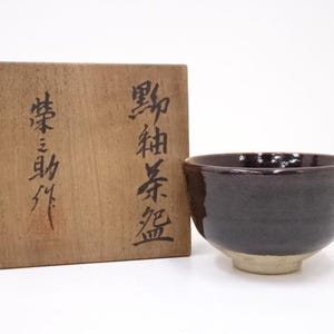May include: A dark brown ceramic tea bowl with a light beige base sits next to a wooden box with Japanese calligraphy. The bowl has a glossy finish and a rounded shape, typical of traditional tea ceremony items.