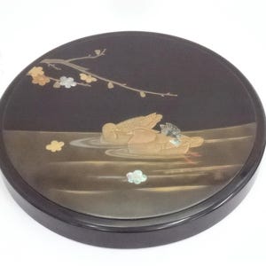 Puede incluir: Bandeja decorativa redonda negra con una ilustración detallada de dos patos en el agua. Los patos son marrones y dorados con detalles azules. Una rama con flores doradas, blancas y plateadas está encima de los patos.